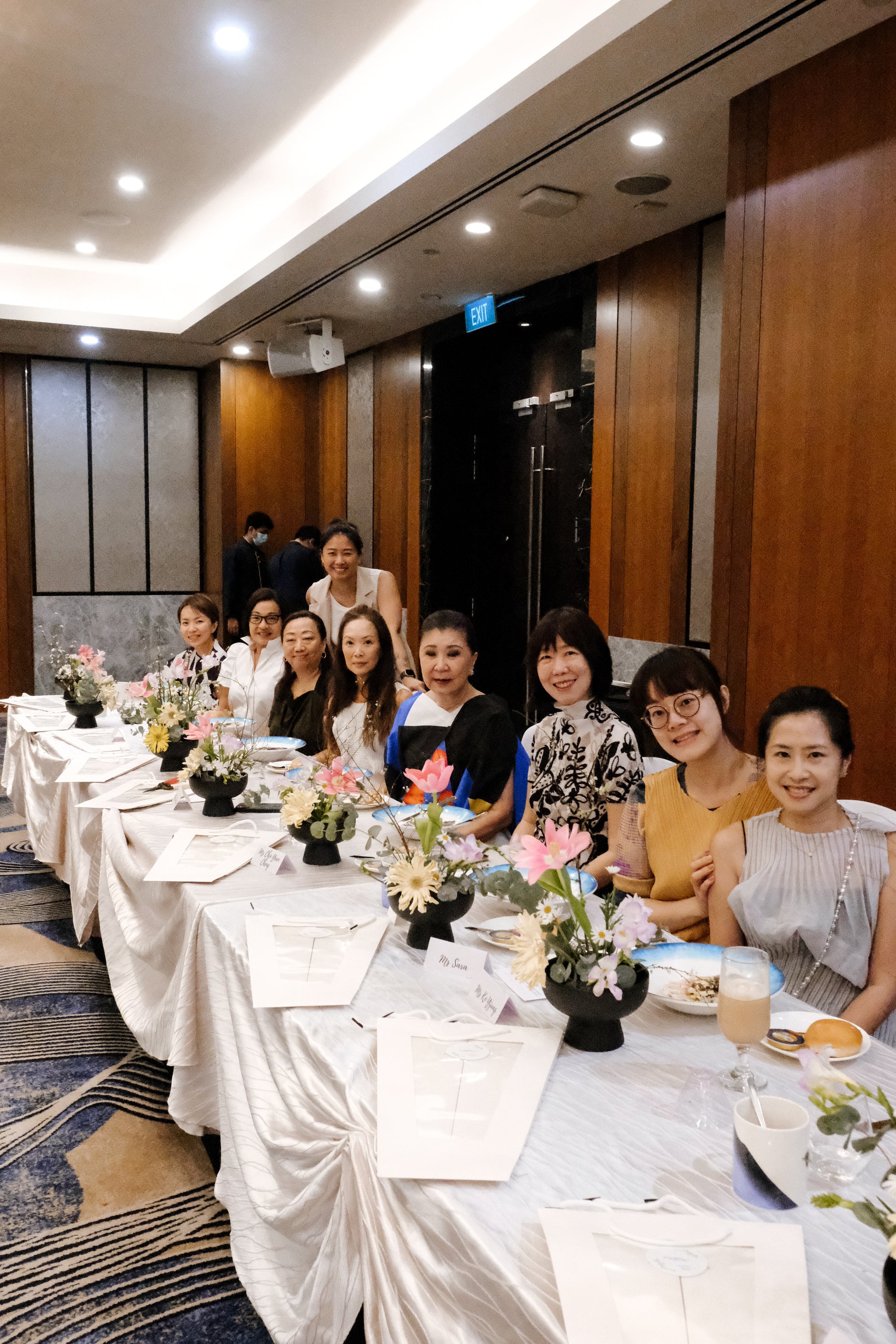 Group of women sitting at a long table with floral arrangements in a formal setting.